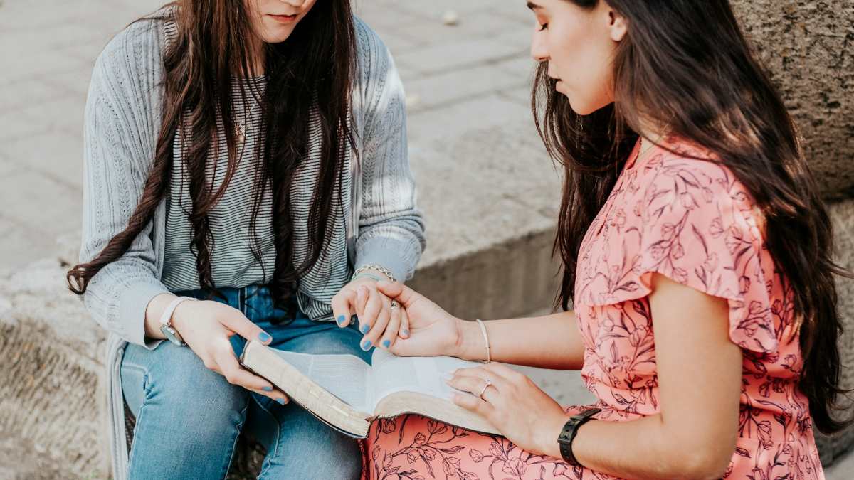 Two women sit side by side sharing and reading a book