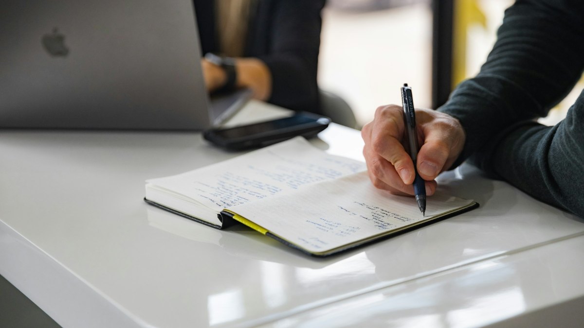 A person writes notes by hand in a notebook beside an open laptop