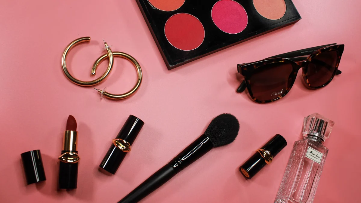 Lipsticks, a makeup palette, earrings and sunglasses on a pink background