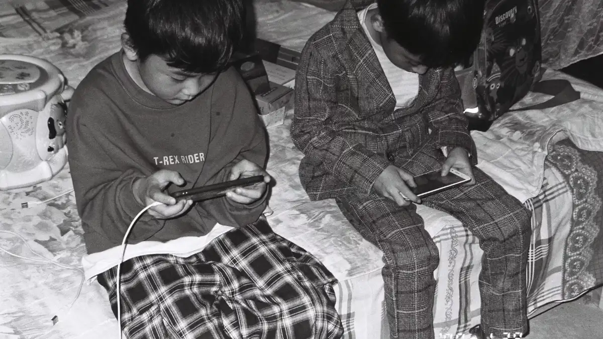 Two boys sitting on a bed looking at their phones